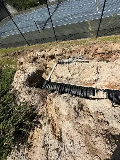 Collapsed dirt slope with exposed drainage pipe beside a chain-link fence and road.