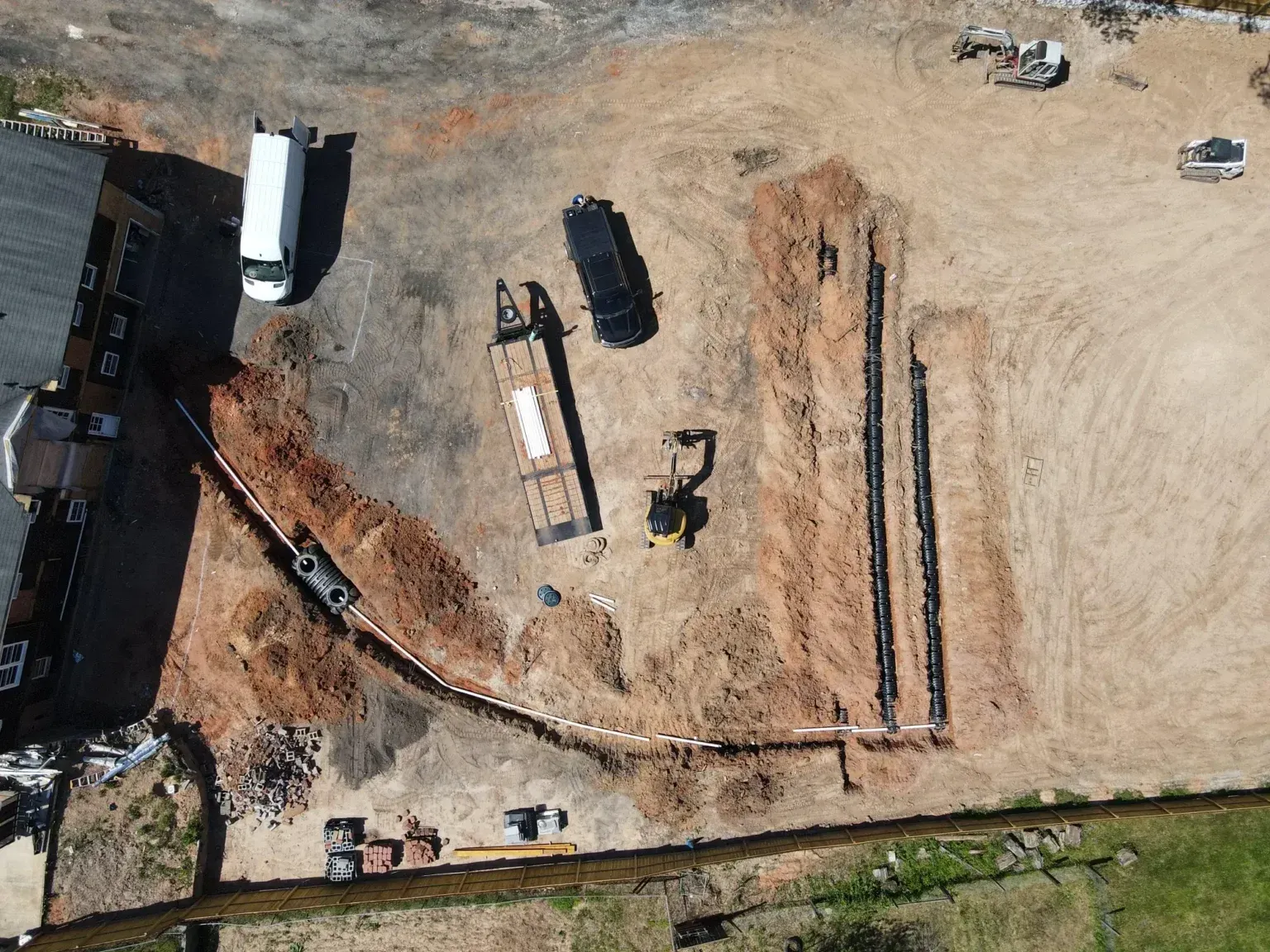Aerial view of a construction site with trucks, equipment, and long trenches on bare dirt.