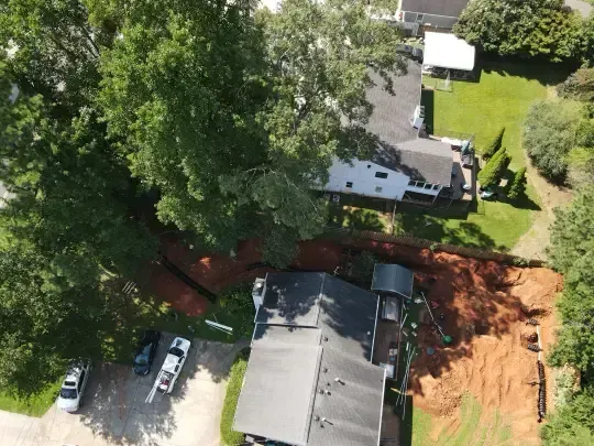 Aerial view of houses, trees, and a dirt excavation area beside a driveway and lawn.