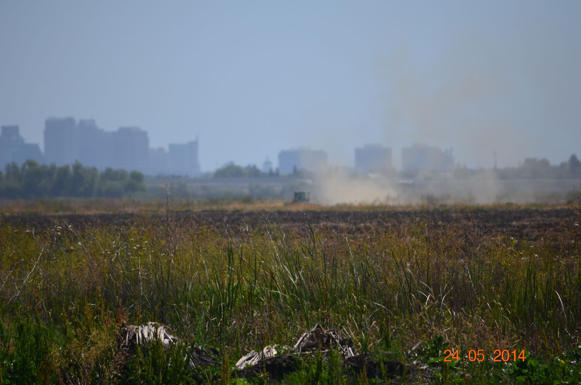 Hazy field with distant city skyline and dust plume under a pale blue sky