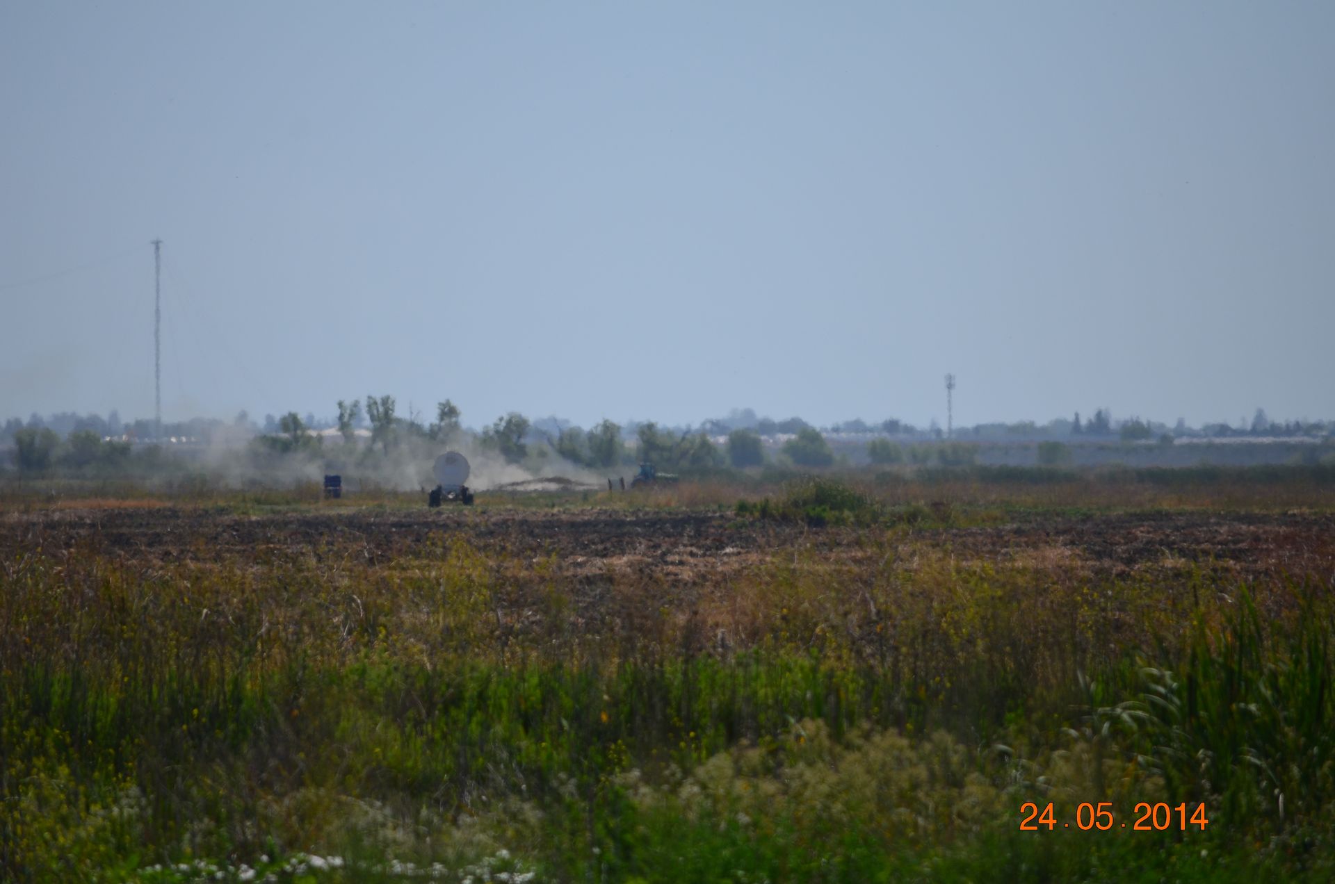 Open field with tall grass and distant trees under a hazy sky, with a few small figures in the distance