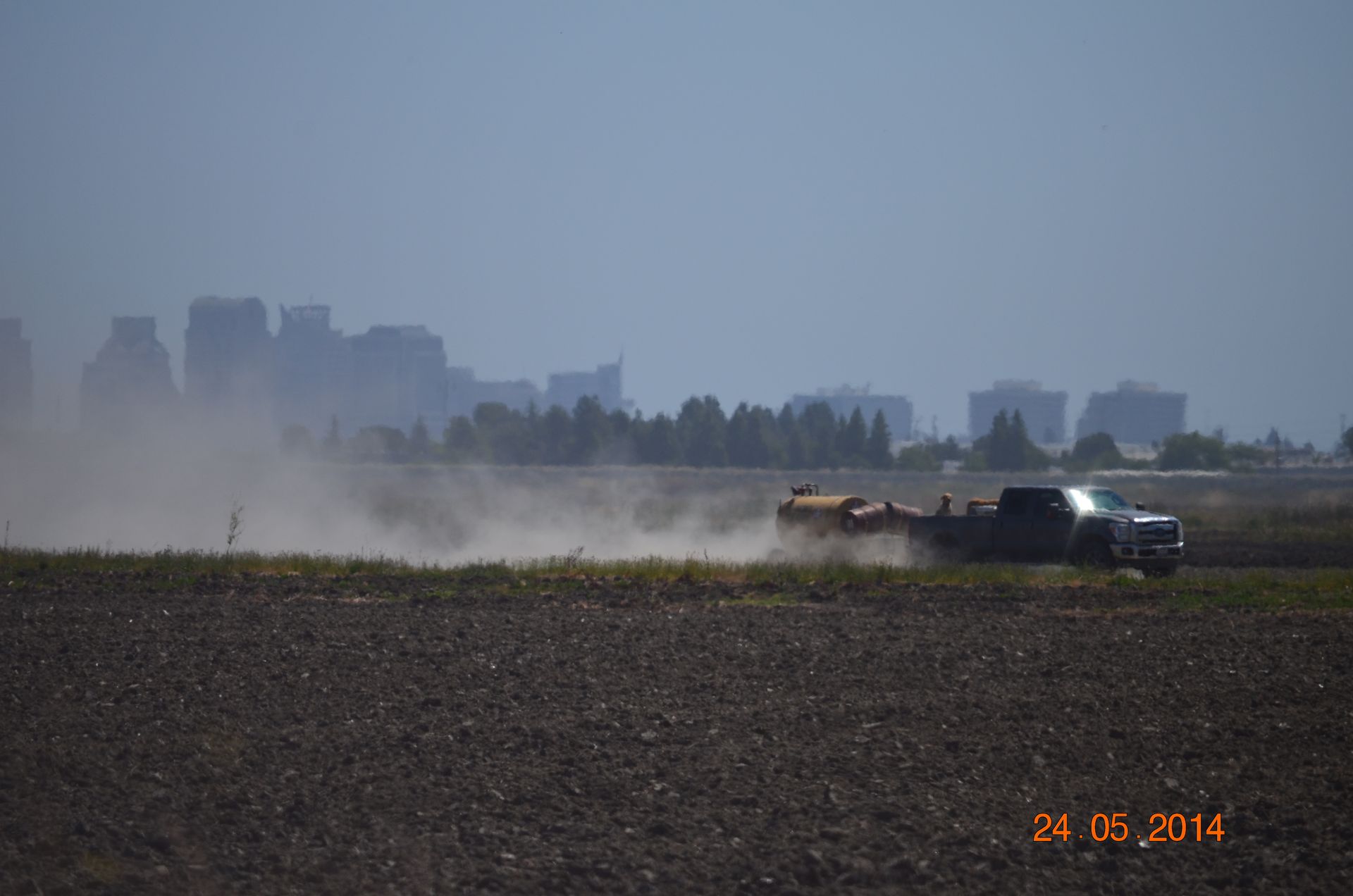Dusty field with a tractor kicking up a cloud of dust, city skyline faintly visible in the background.