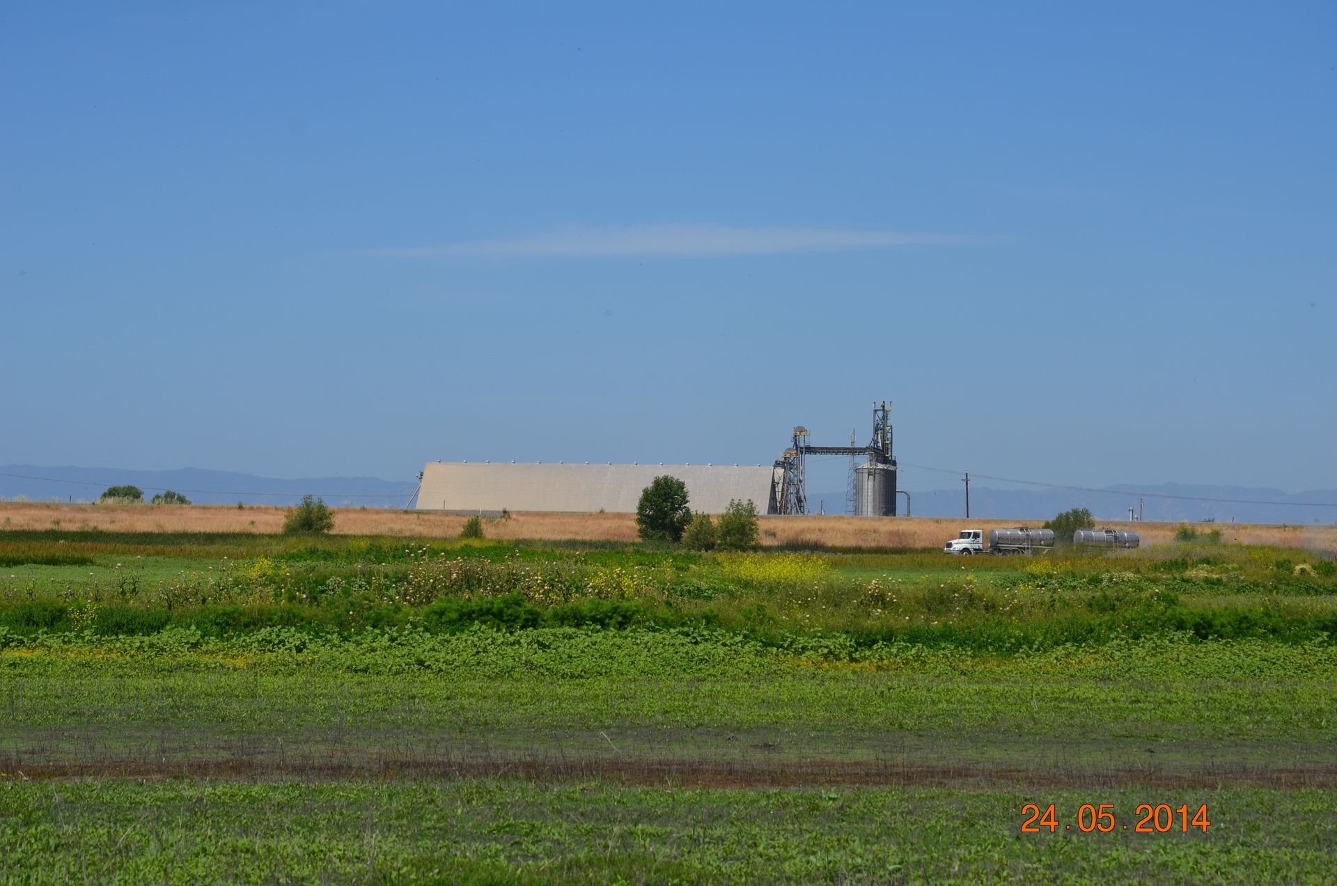 Rural industrial site with grain silos and a tank beside green fields under a clear blue sky