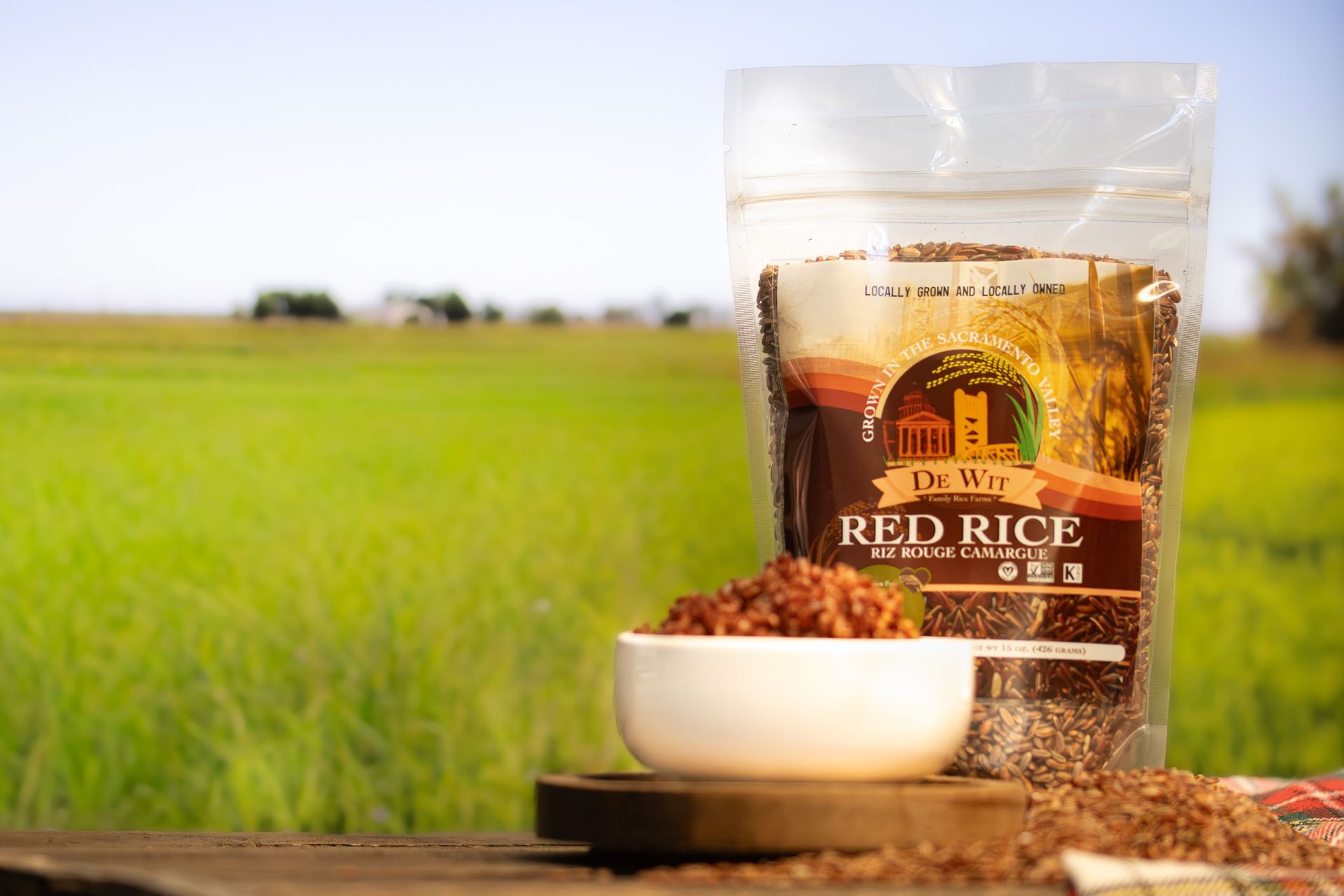 Bag of DeWit Red Rouge Camargue beside a bowl of rice on a wooden table with a sunlit field background