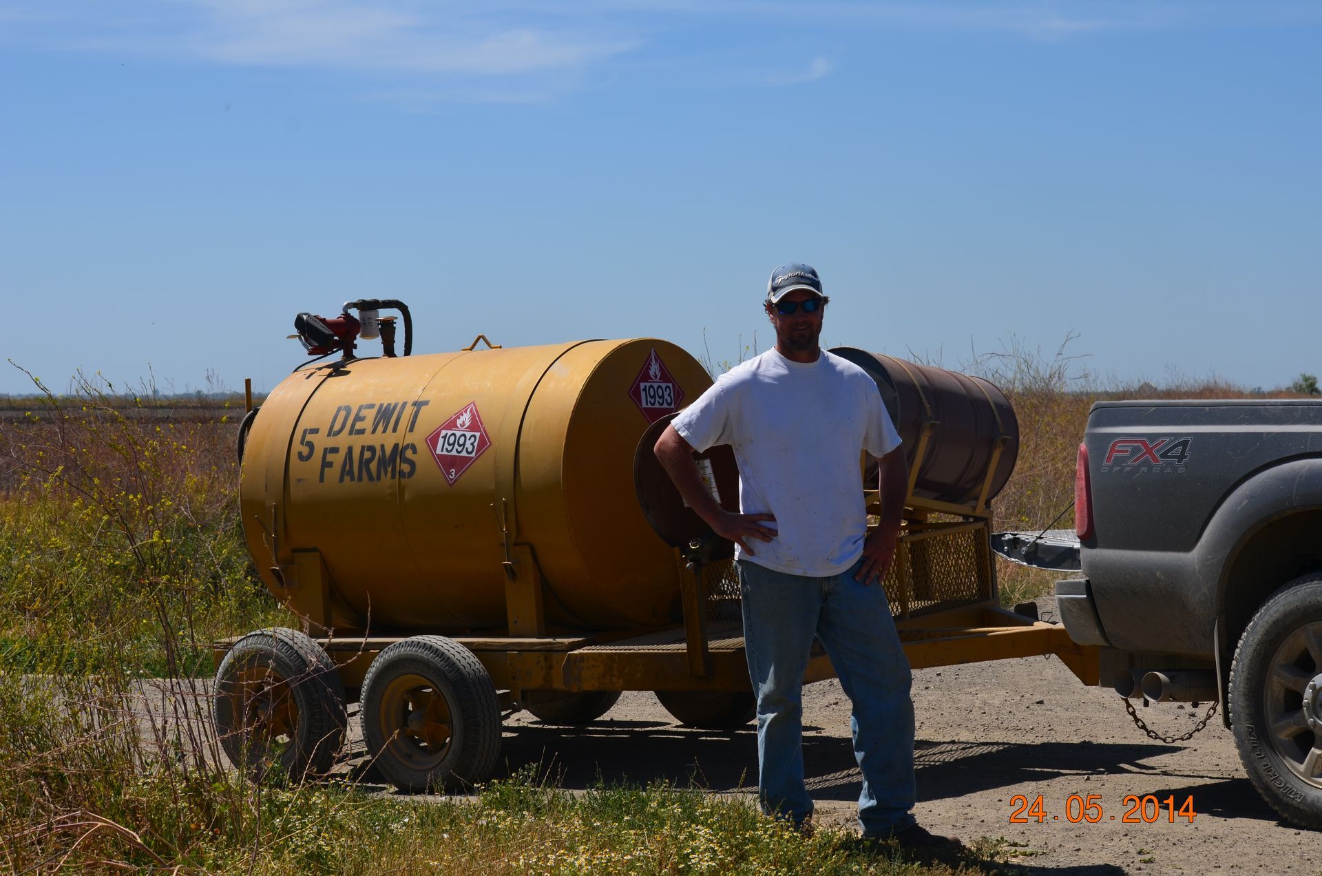Man standing beside a yellow farm sprayer hitched to a pickup on a rural roadside