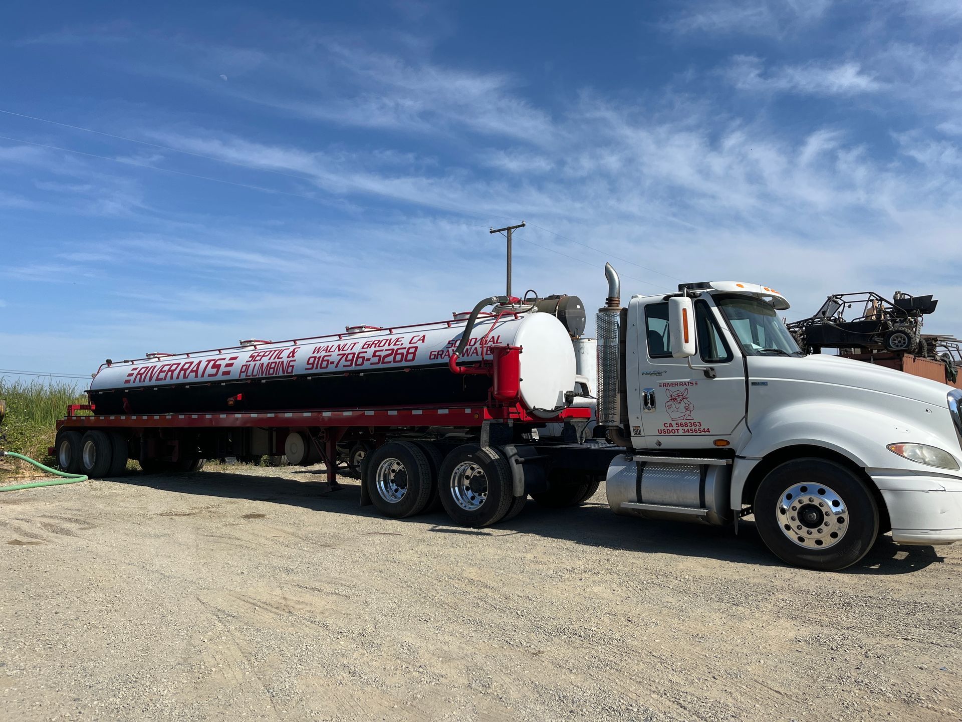 White semi truck hauling a long black tanker trailer on a gravel road under a blue sky