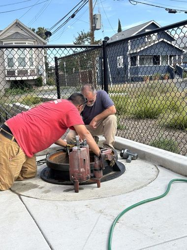 Two workers service a pump inside a fenced utility pit beside a house.