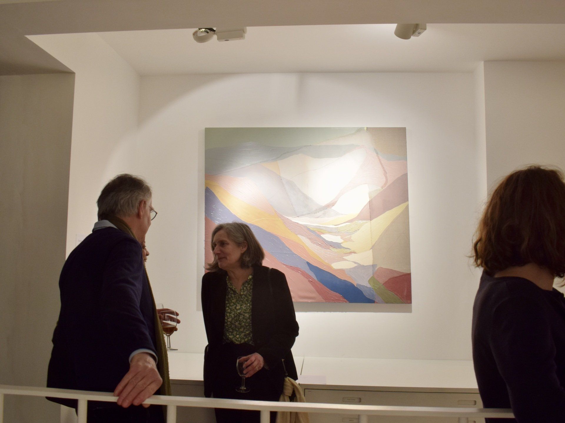 An abstract painting on a white wall with two people standing talking in front