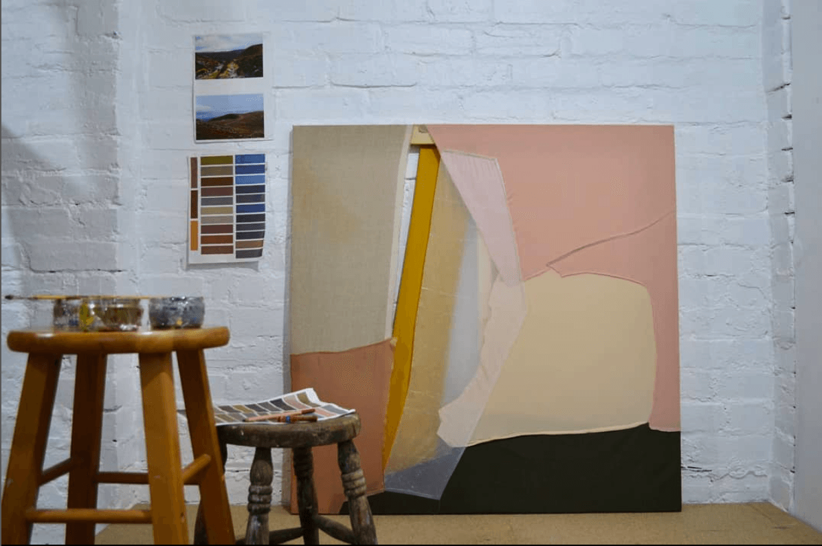 Photo of artist studio with white walls and two stools. A pink and yellow painting is leaning against the wall