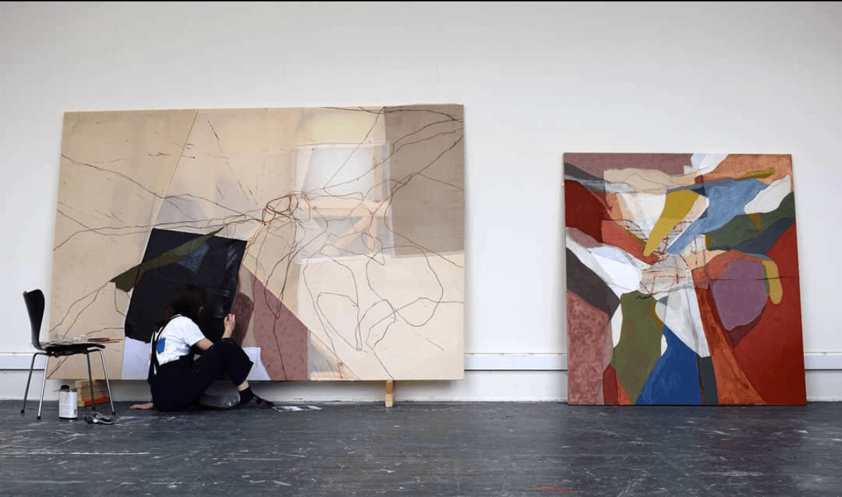Photo of artist crouched, painting on large canvas against a white studio wall