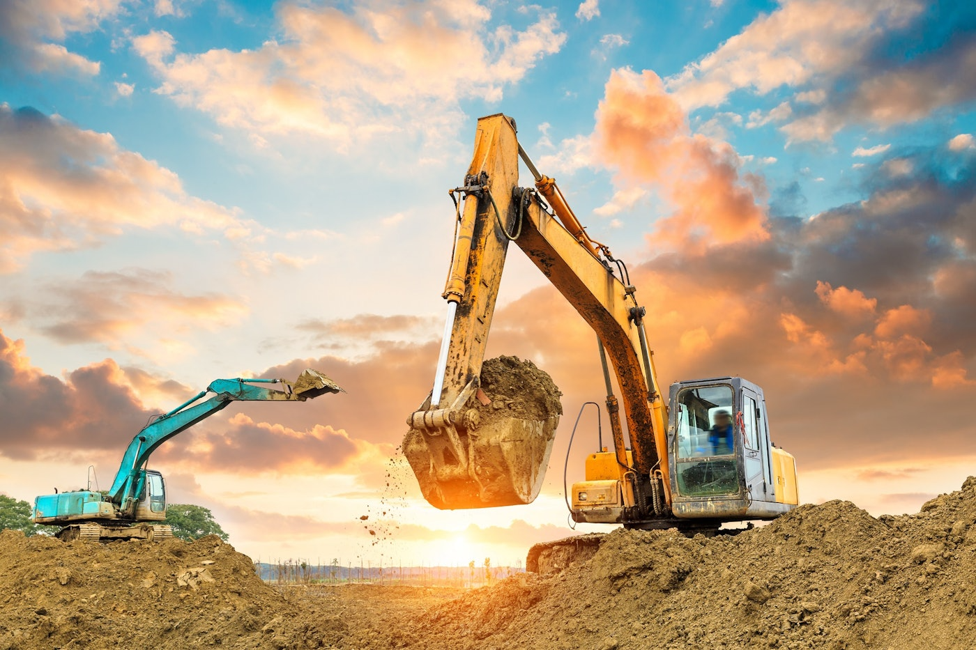 Two excavators digging earth on a construction site; blue and yellow machinery against a sunset.