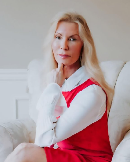 Woman in red dress and white shirt, sitting in an armchair, touching her neck, looking at the camera.