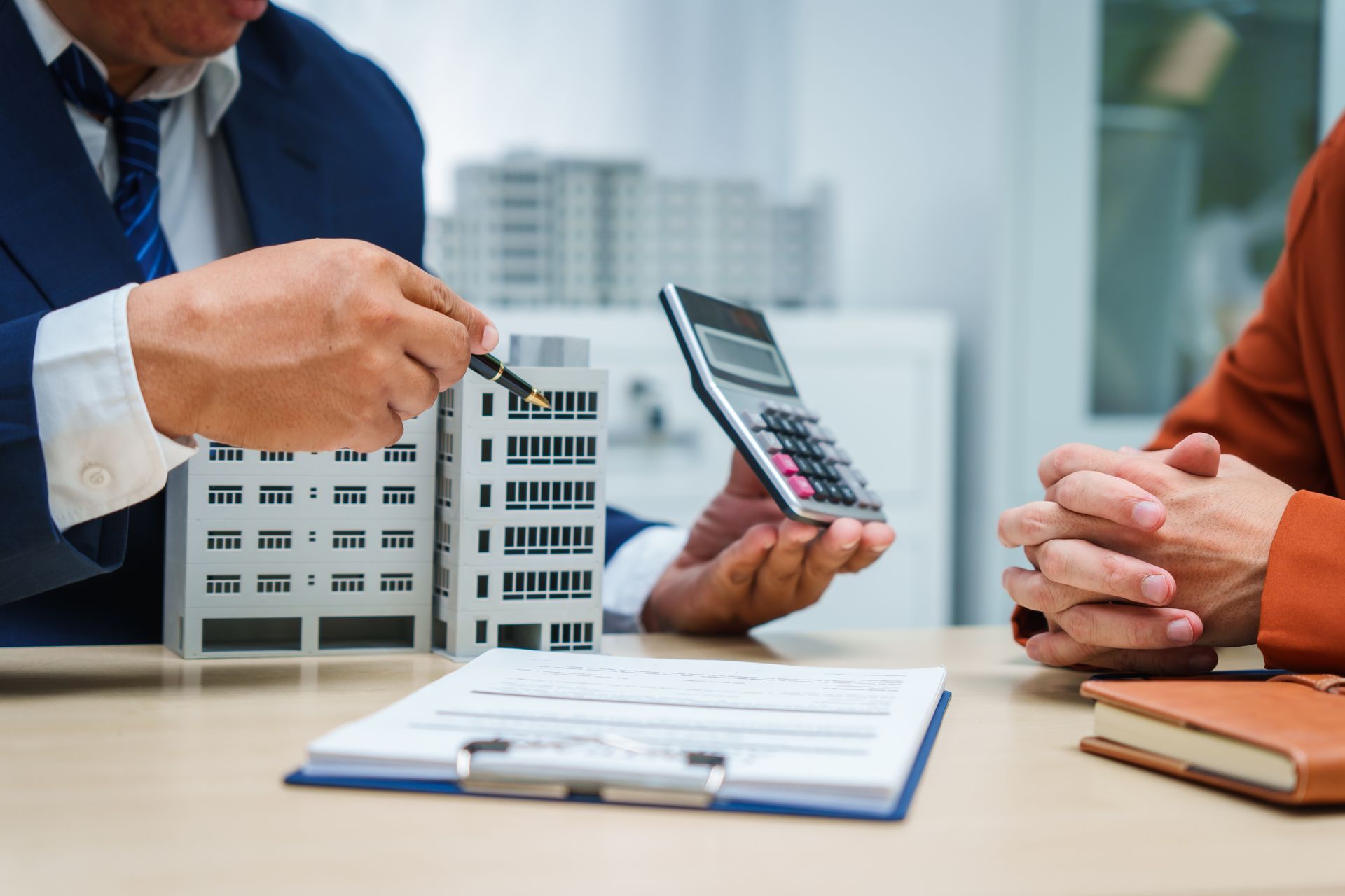 Real estate agent pointing at model building, reviewing paperwork with client, calculator visible.