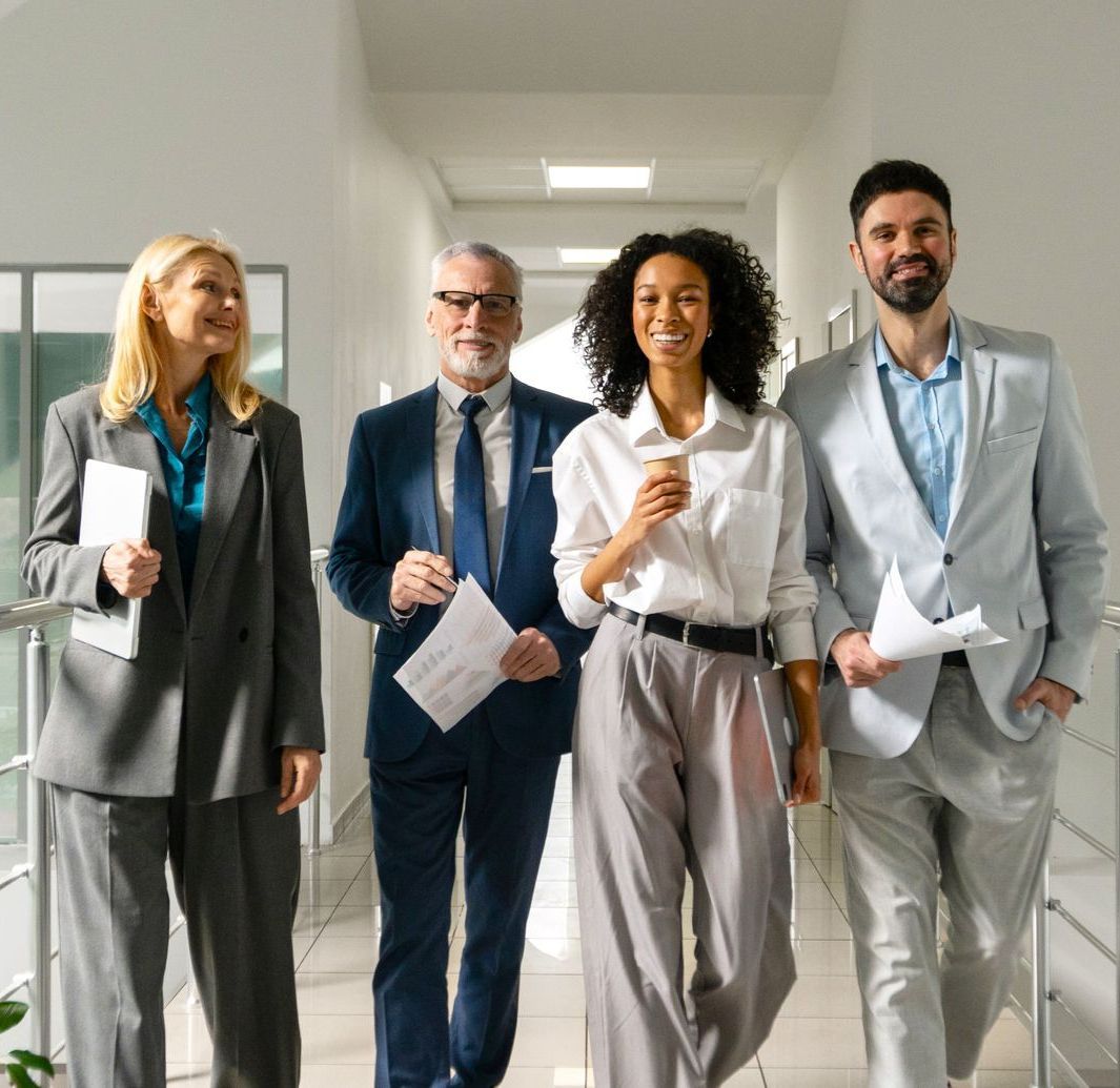 Four people walking down a bright hallway, smiling, holding papers, and dressed in business attire.