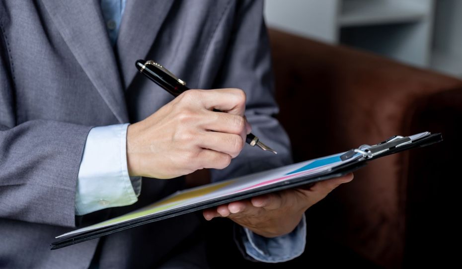 Person in a suit holding a pen and clipboard, about to write.