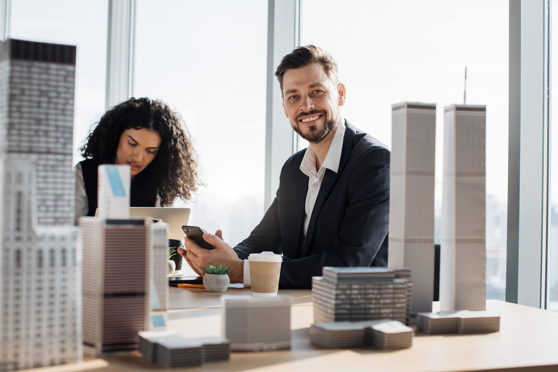 Man smiling, holding phone, at table with woman, model buildings, coffee cup, sunny office setting.