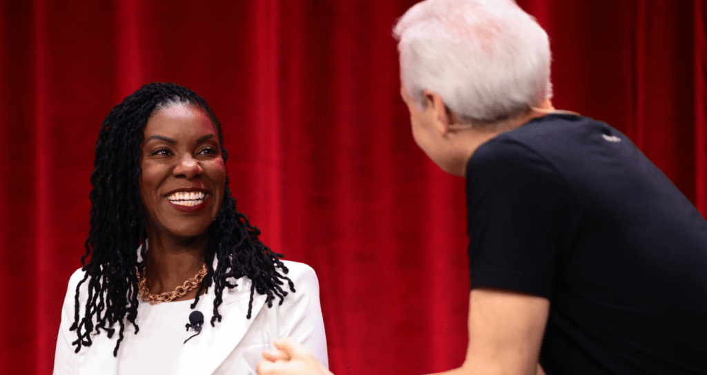 Woman in white blazer smiles, conversing with a man in black t-shirt near red curtains.
