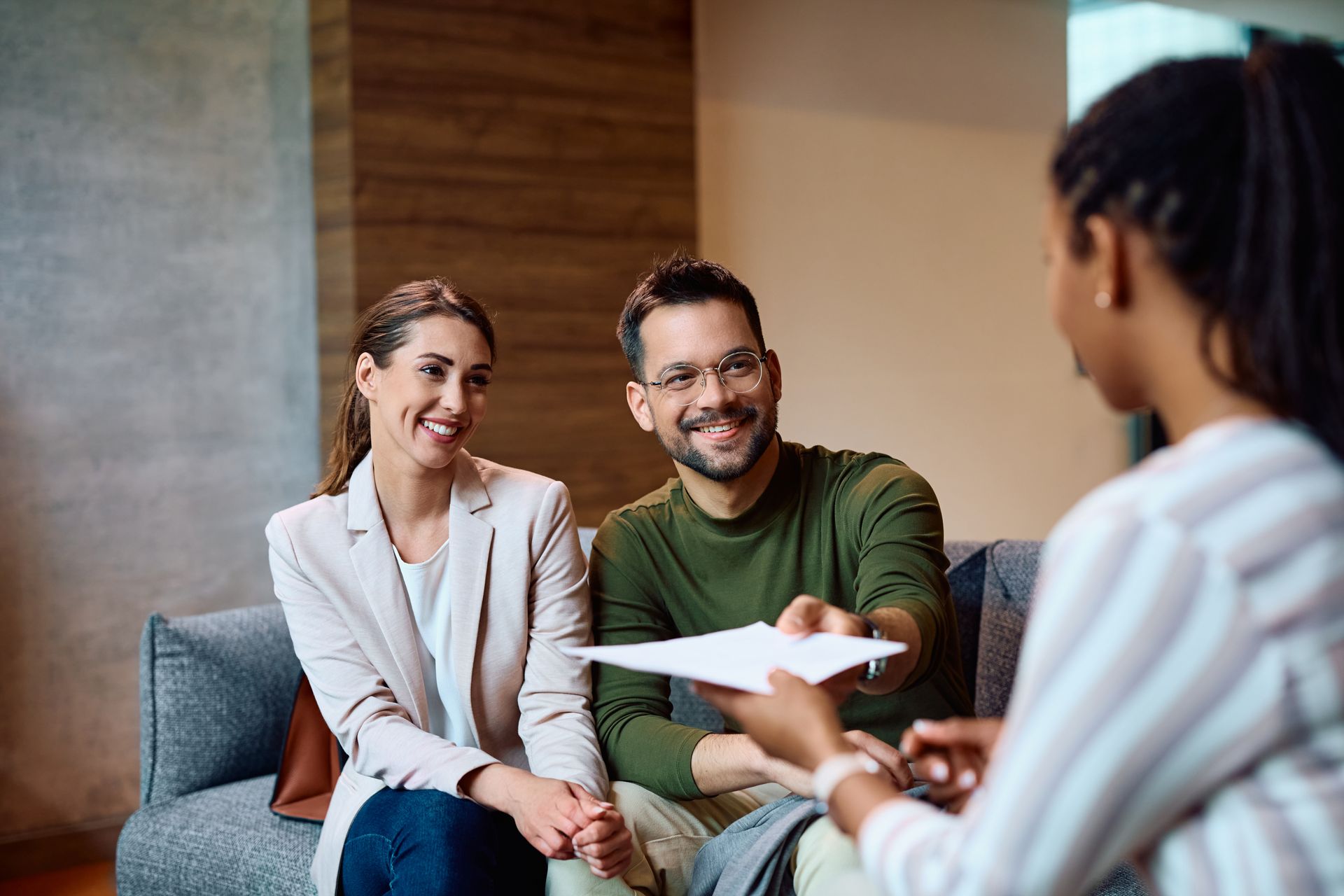 A couple smiles as someone hands them paperwork, indoors on a sofa.