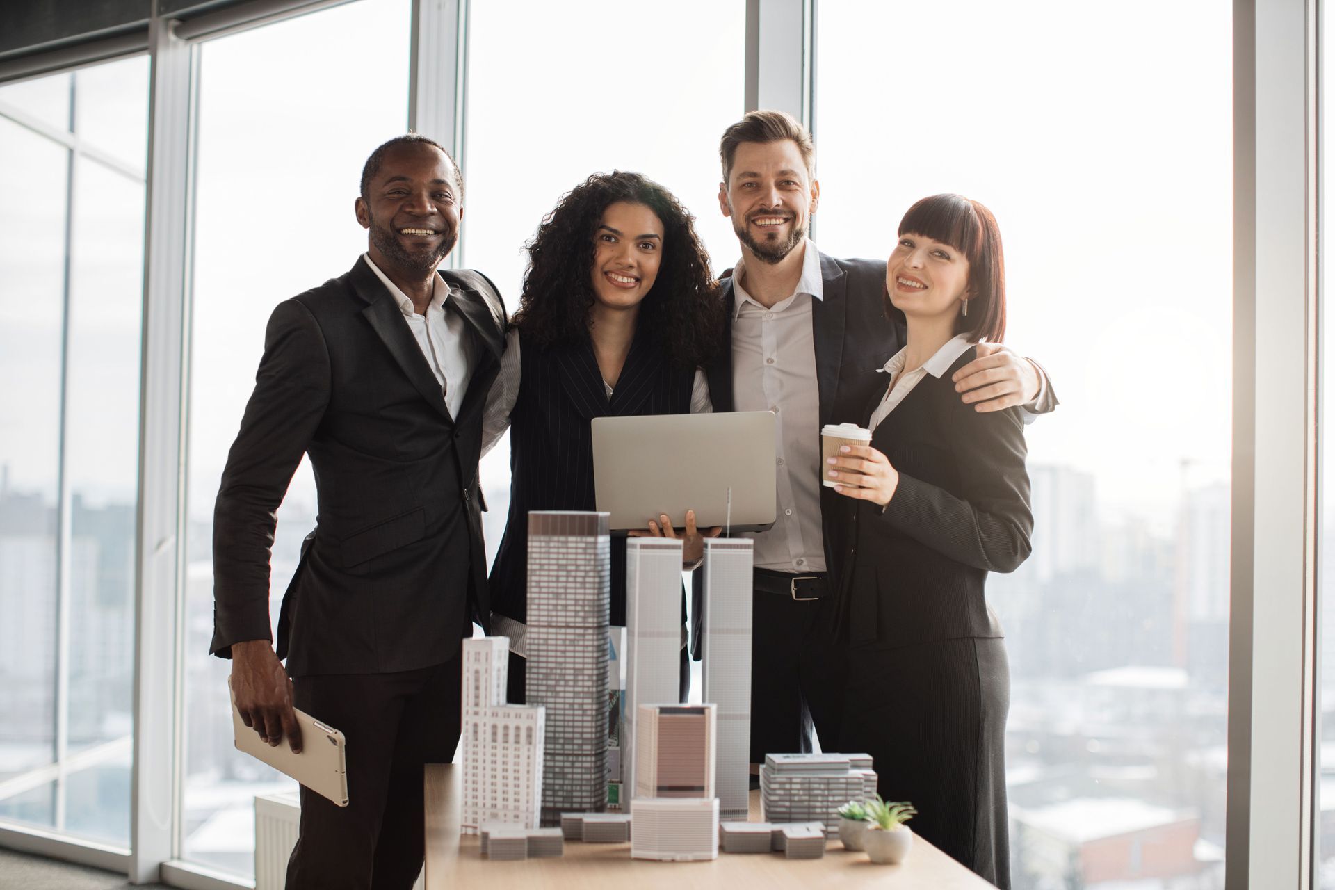 Four businesspeople stand together, smiling, with building models and laptop on table near a window.