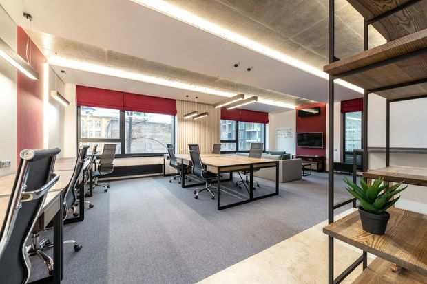 Modern office space with desks, chairs, windows, and shelving unit. Neutral colors and soft lighting.