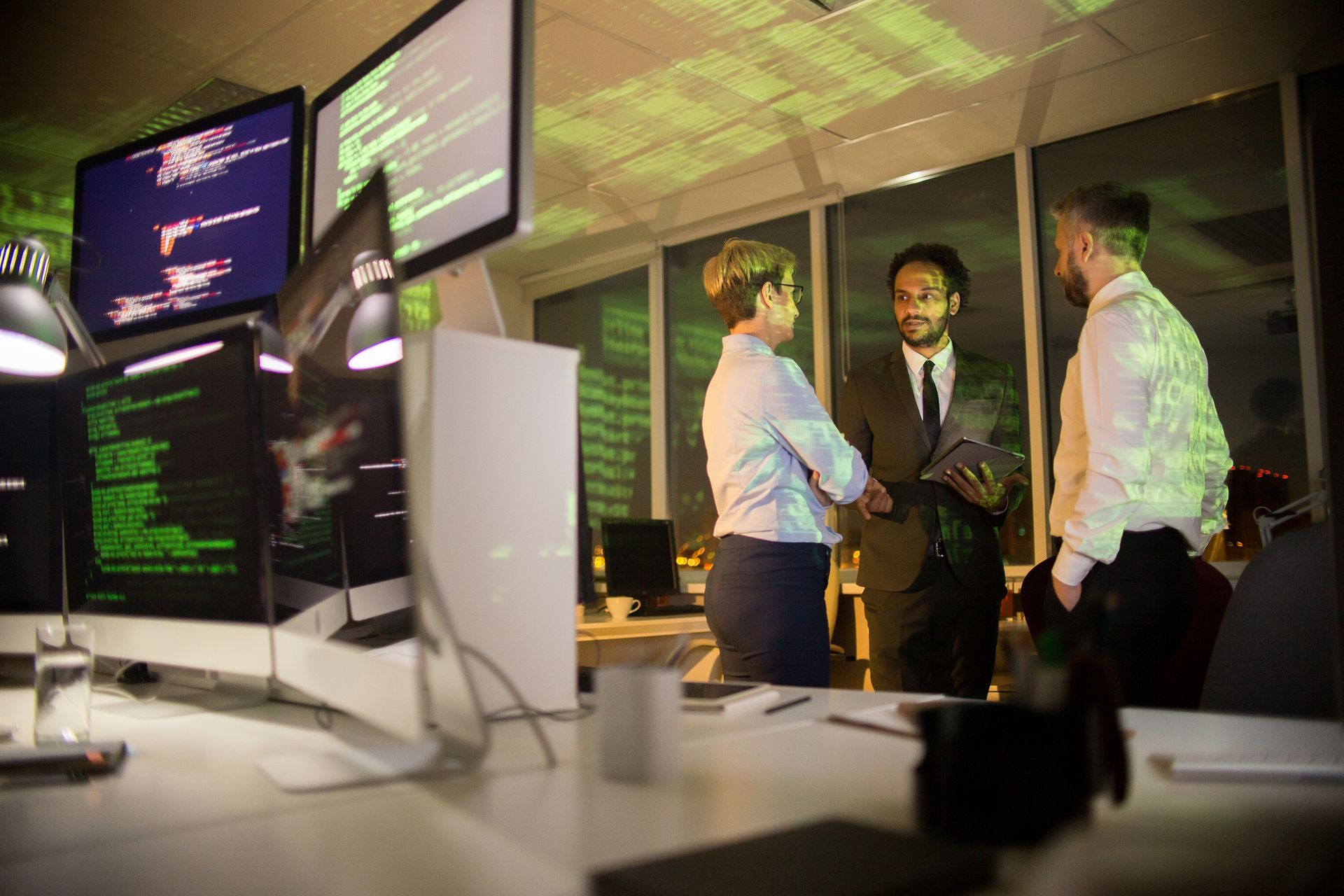 Three people in an office with computer screens displaying code, discussing, with city lights visible outside.