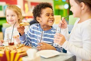 Three children laughing and eating pizza together at a brightly colored indoor venue.