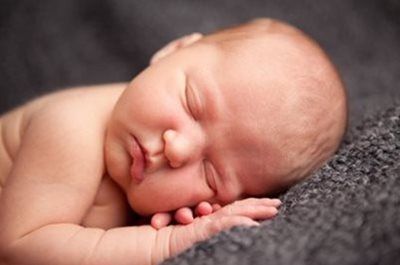 A newborn sleeping peacefully with their cheek resting on their small hands against a textured grey background.