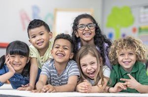 Six diverse children lying on their stomachs, smiling and laughing together in a brightly decorated classroom.