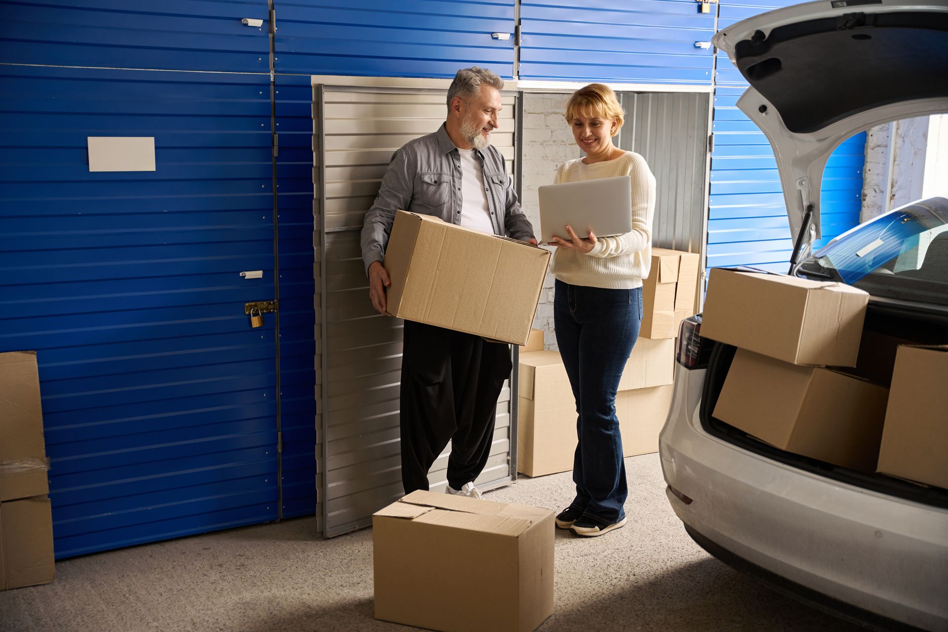 Man and woman loading cardboard boxes into a car trunk outside a budget-friendly storage unit. Man and woman loading cardboard boxes into a car trunk outside a budget-friendly storage unit.