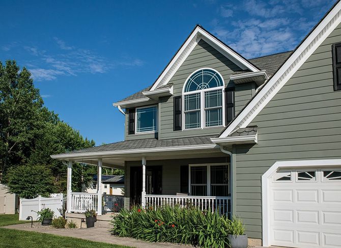 A two-story green house with a front porch, white railings, and an arched window under a bright blue sky.