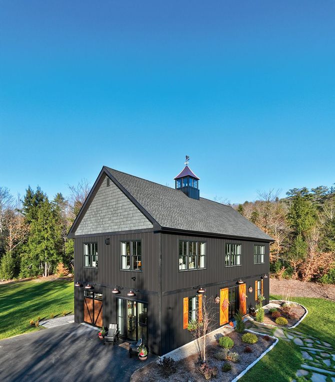 A black, two-story barn-style building with a cupola stands on a grassy lot next to a paved driveway and stone path.