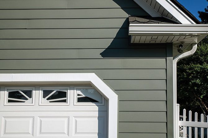 An exterior view of a garage with sage green horizontal siding, a white door with rectangular windows, and a white fence.