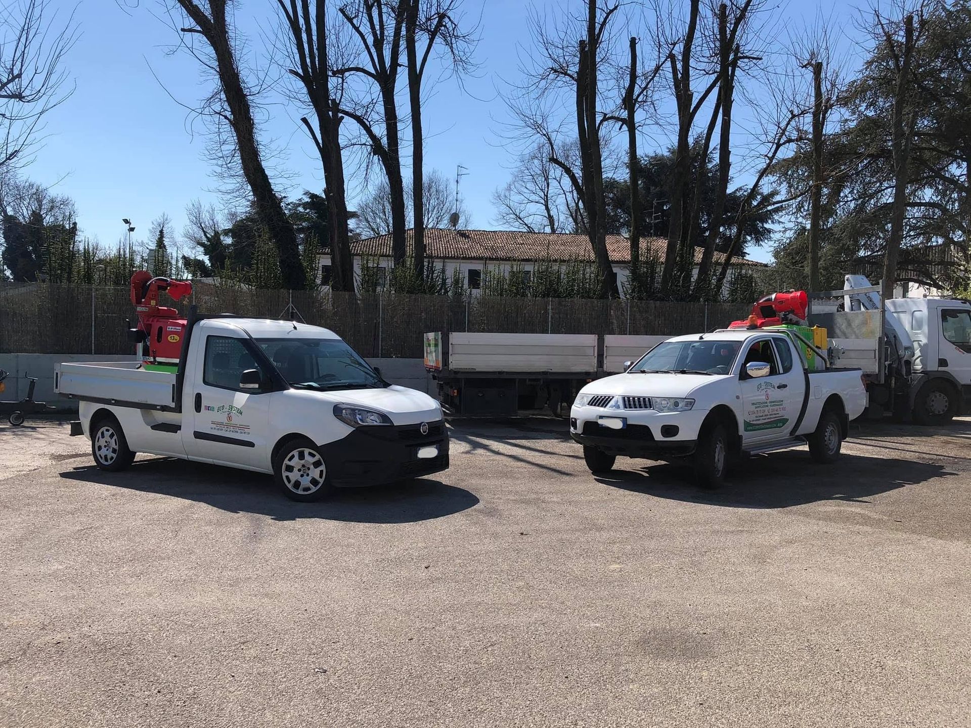Due pick-up bianchi da lavoro parcheggiati su ghiaia in un piazzale, con alberi e una recinzione sullo sfondo.