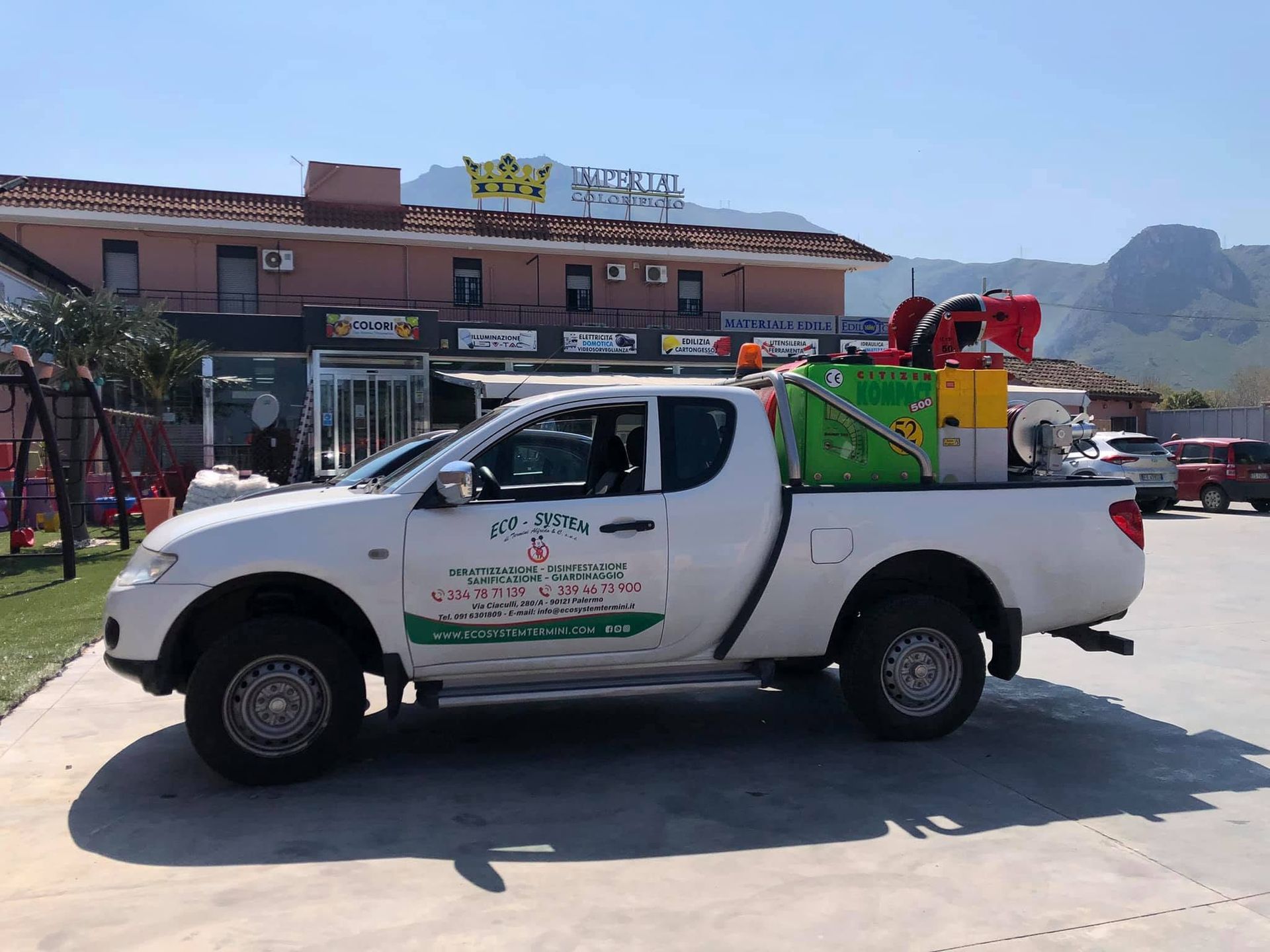 Un pick-up bianco carico di provviste è parcheggiato fuori da un edificio, con le montagne sullo sfondo.