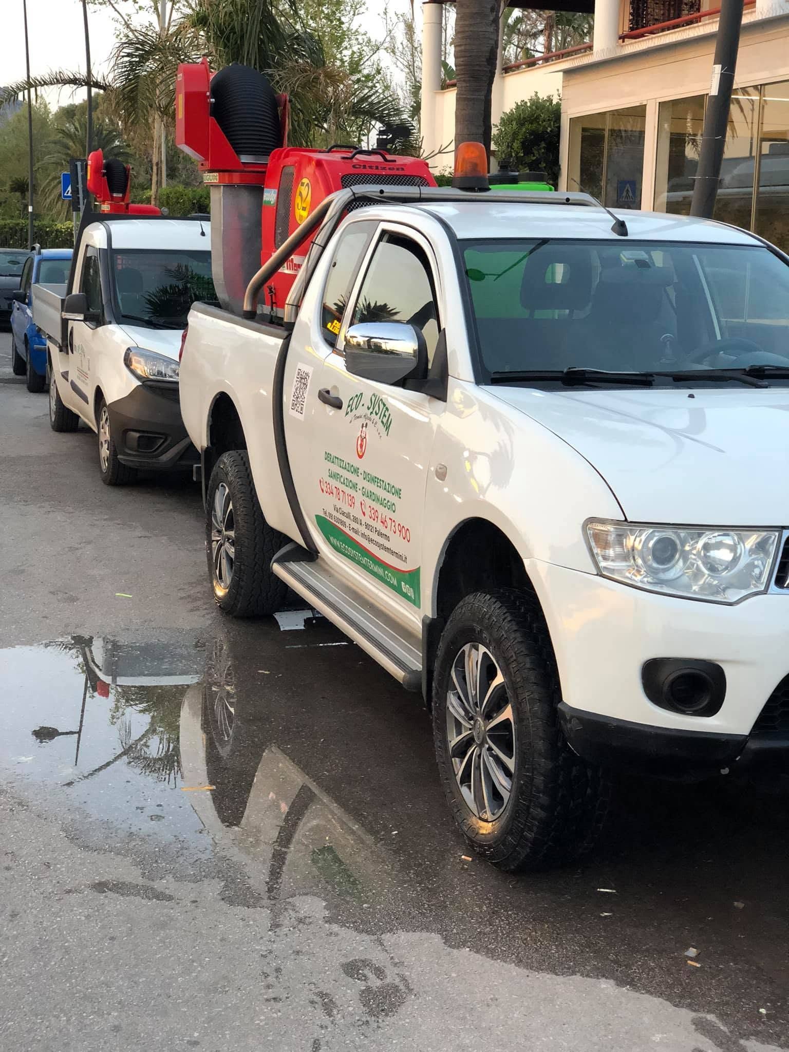 Due pick-up bianchi parcheggiati in strada, uno con un carrello elevatore rosso montato sul cassone, accanto a una pozzanghera.
