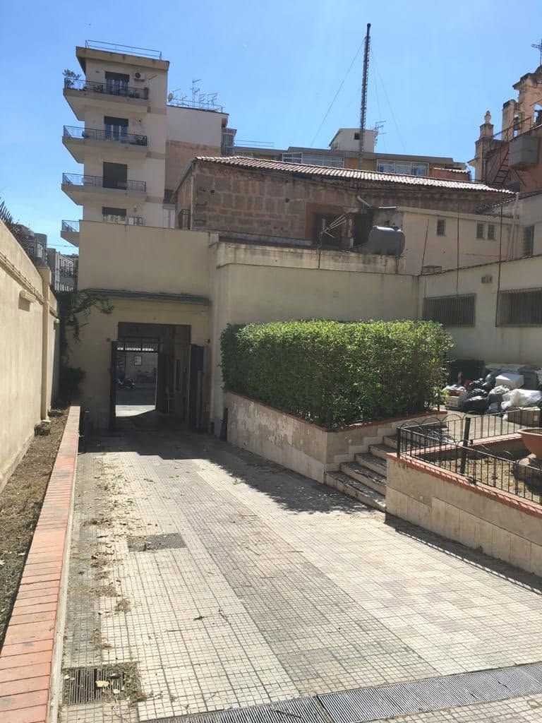 Stretto vicolo soleggiato con pavimentazione piastrellata, palazzi e una siepe accanto a un piccolo passaggio ad arco.