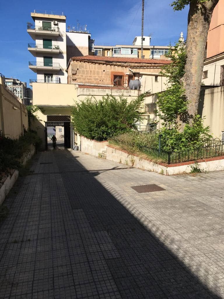 Stretto vicolo soleggiato tra gli edifici, con un marciapiede lastricato e alberi, che conduce a una piccola via cittadina.