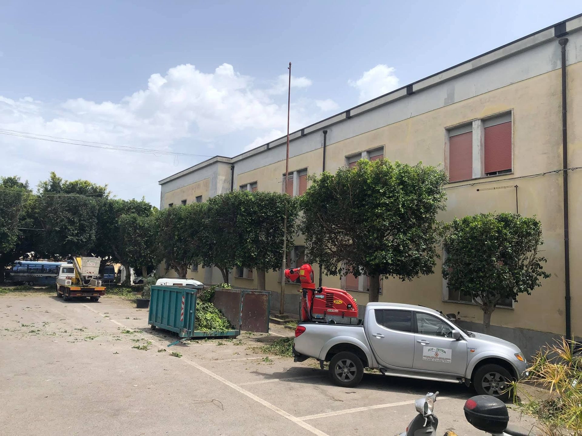 Edificio industriale con piccoli alberi, un pick-up parcheggiato, un cassonetto per i rifiuti e macchinari rossi in un lotto soleggiato.