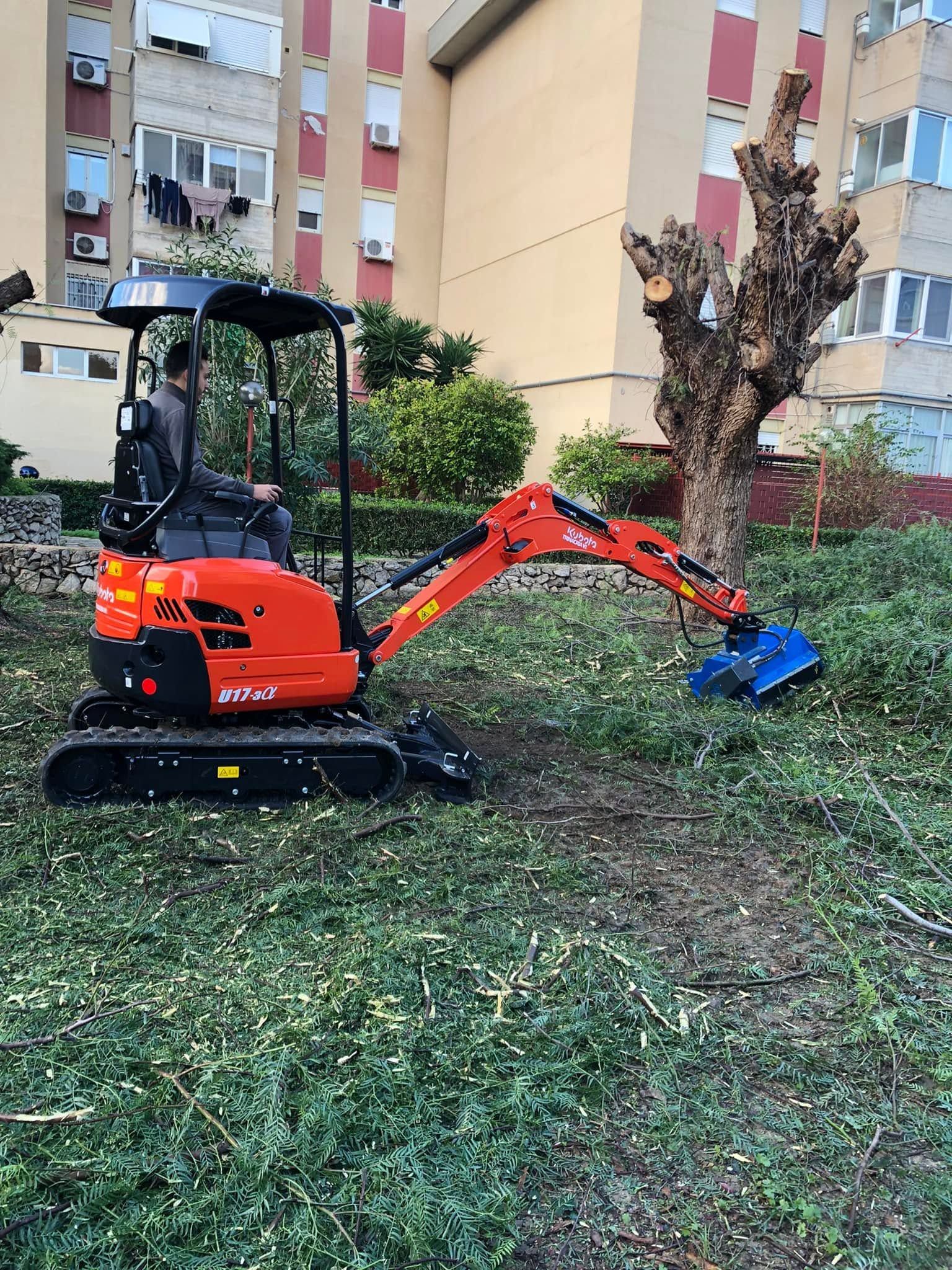 Piccolo escavatore arancione con benna blu che ripulisce l'erba vicino a un condominio.