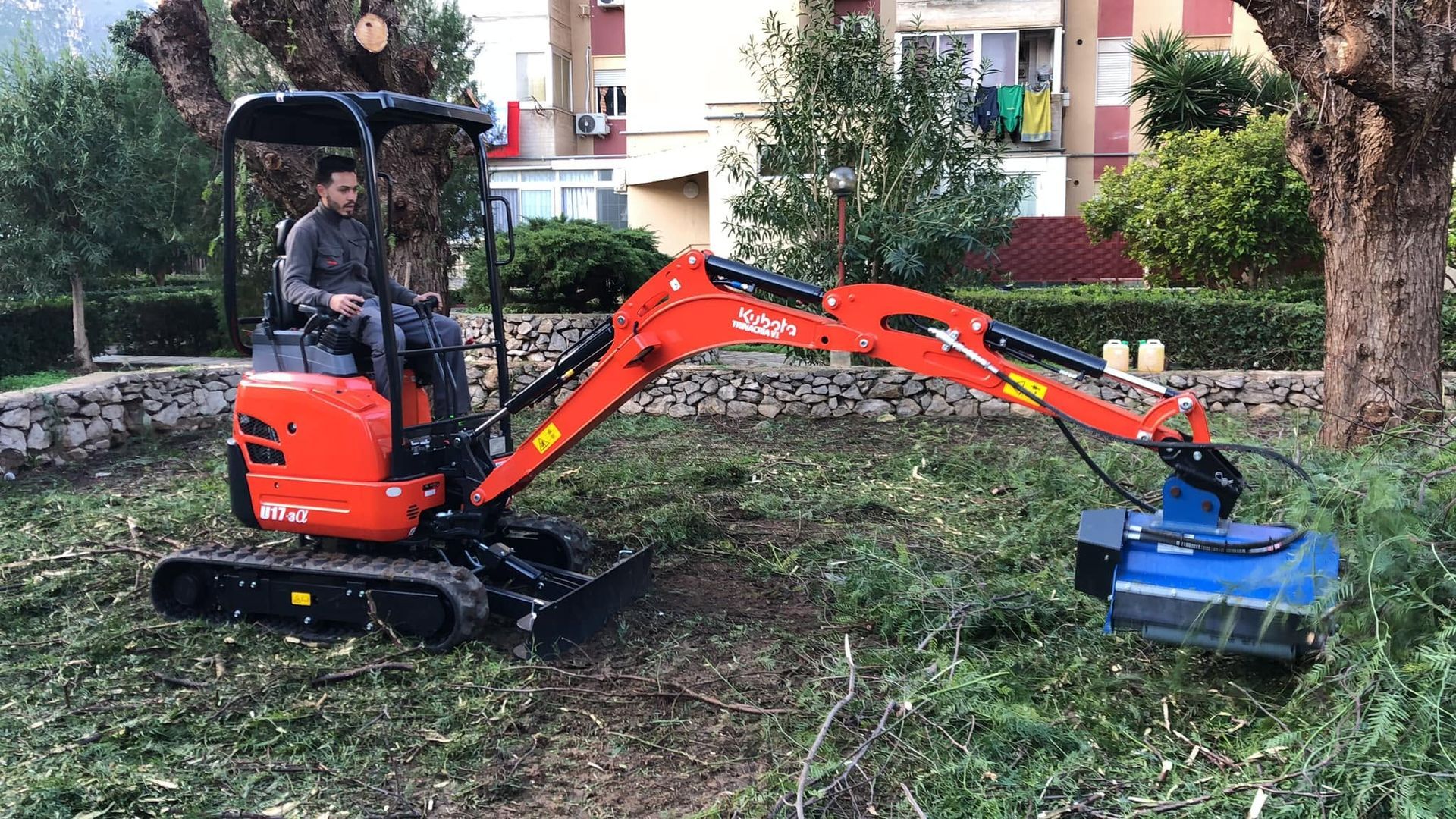 Mini escavatore arancione che falcia l'erba alta in un cortile con un accessorio blu.
