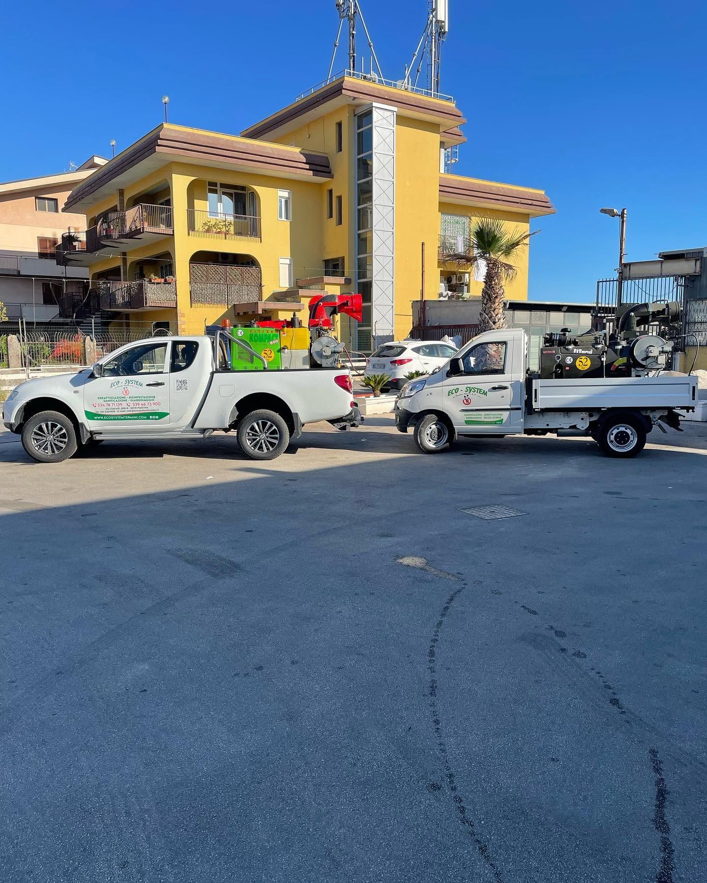 Camion di servizio bianchi parcheggiati fuori da un edificio giallo a due piani, con attrezzature edili e gru nelle vicinanze.