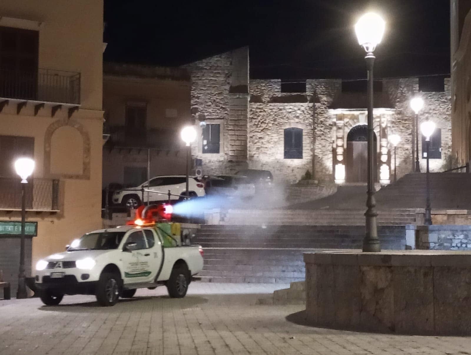 Un pick-up bianco spruzza nebbia in una piazza di pietra illuminata di notte, con edifici e lampioni sullo sfondo.