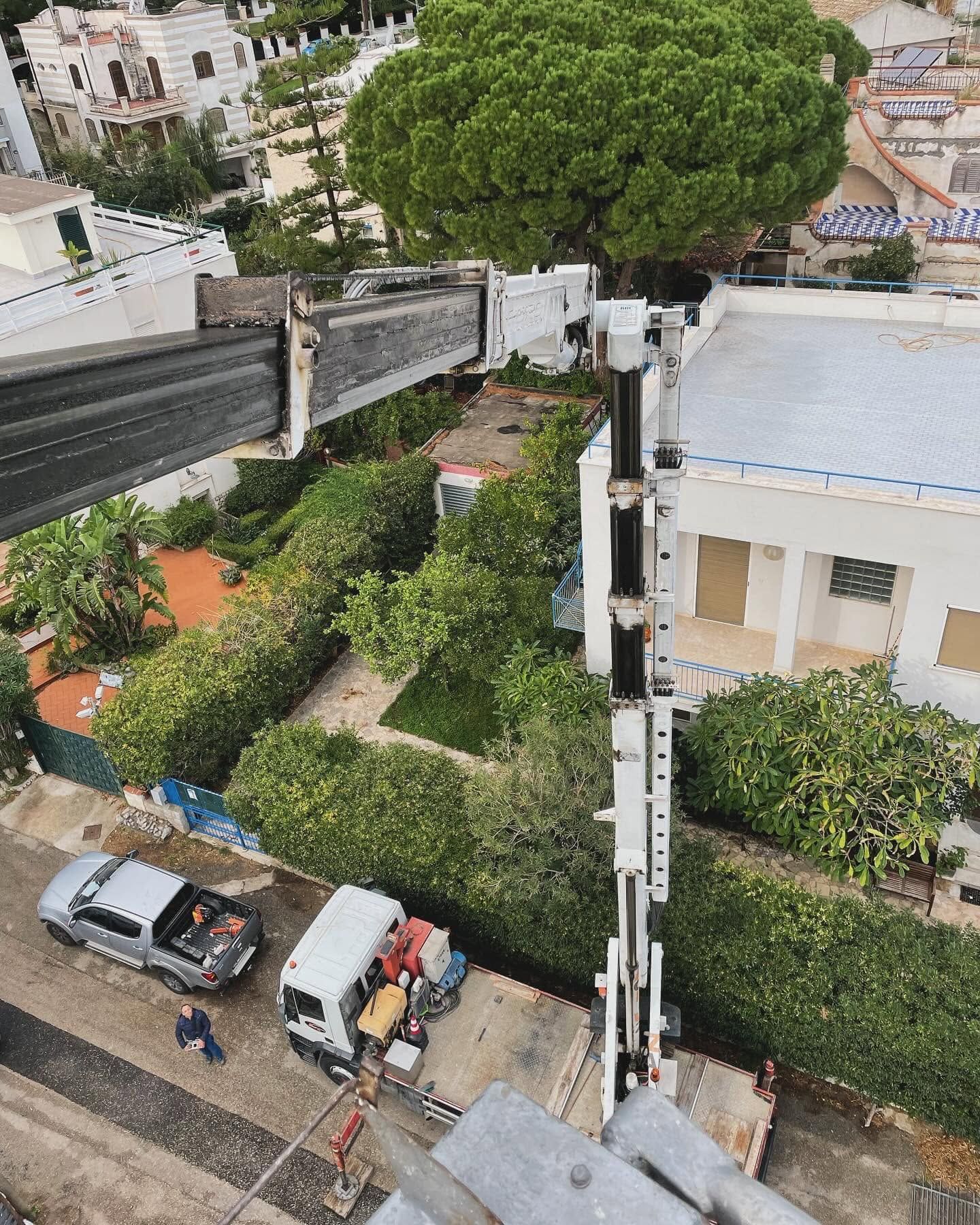 Veduta aerea di una strada residenziale con case, alberi, veicoli parcheggiati e un grande braccio di gru in alto.
