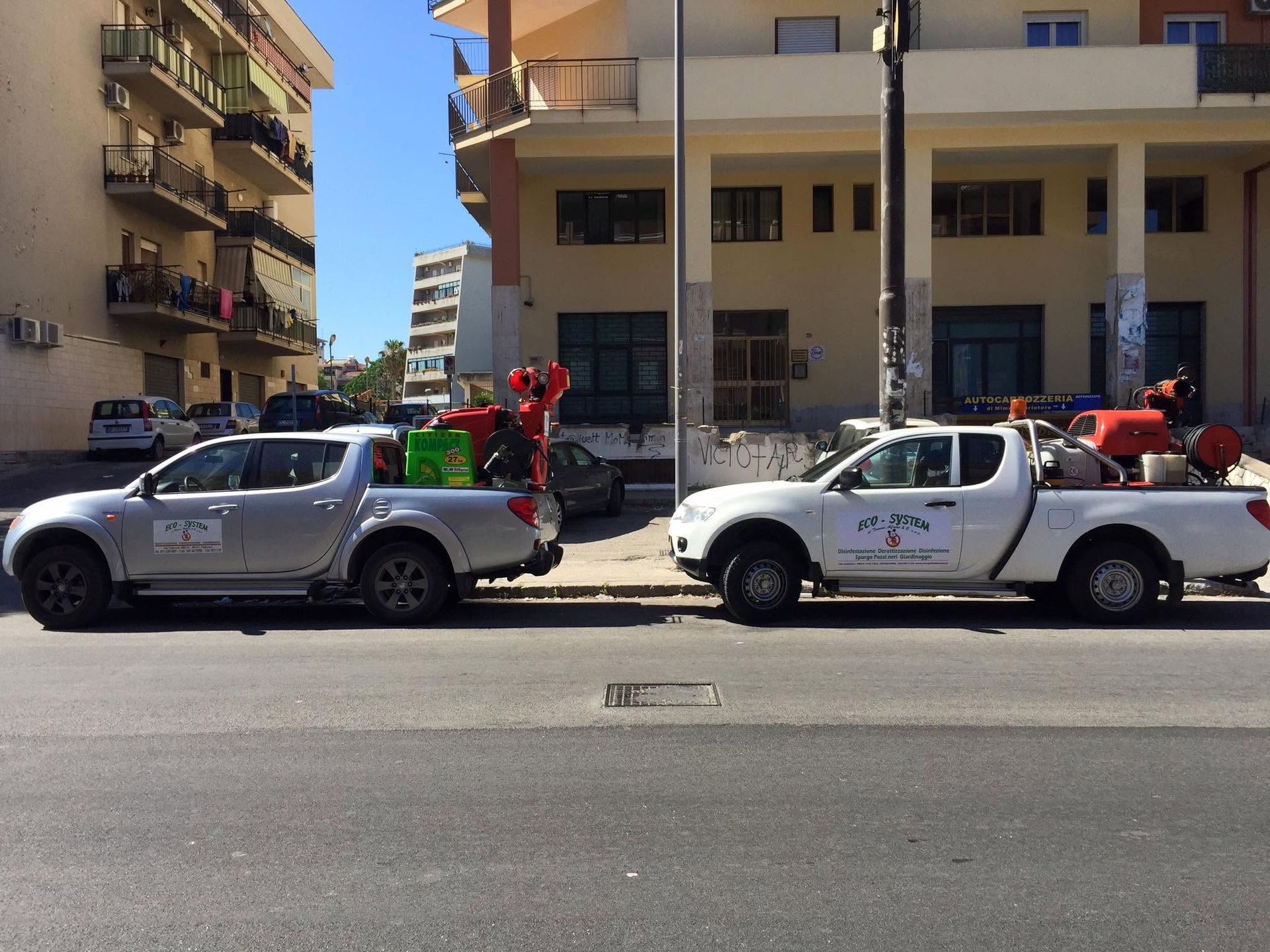 Due pick-up con persone e bandiere a bordo, parcheggiati su una strada cittadina accanto ad alcuni edifici.