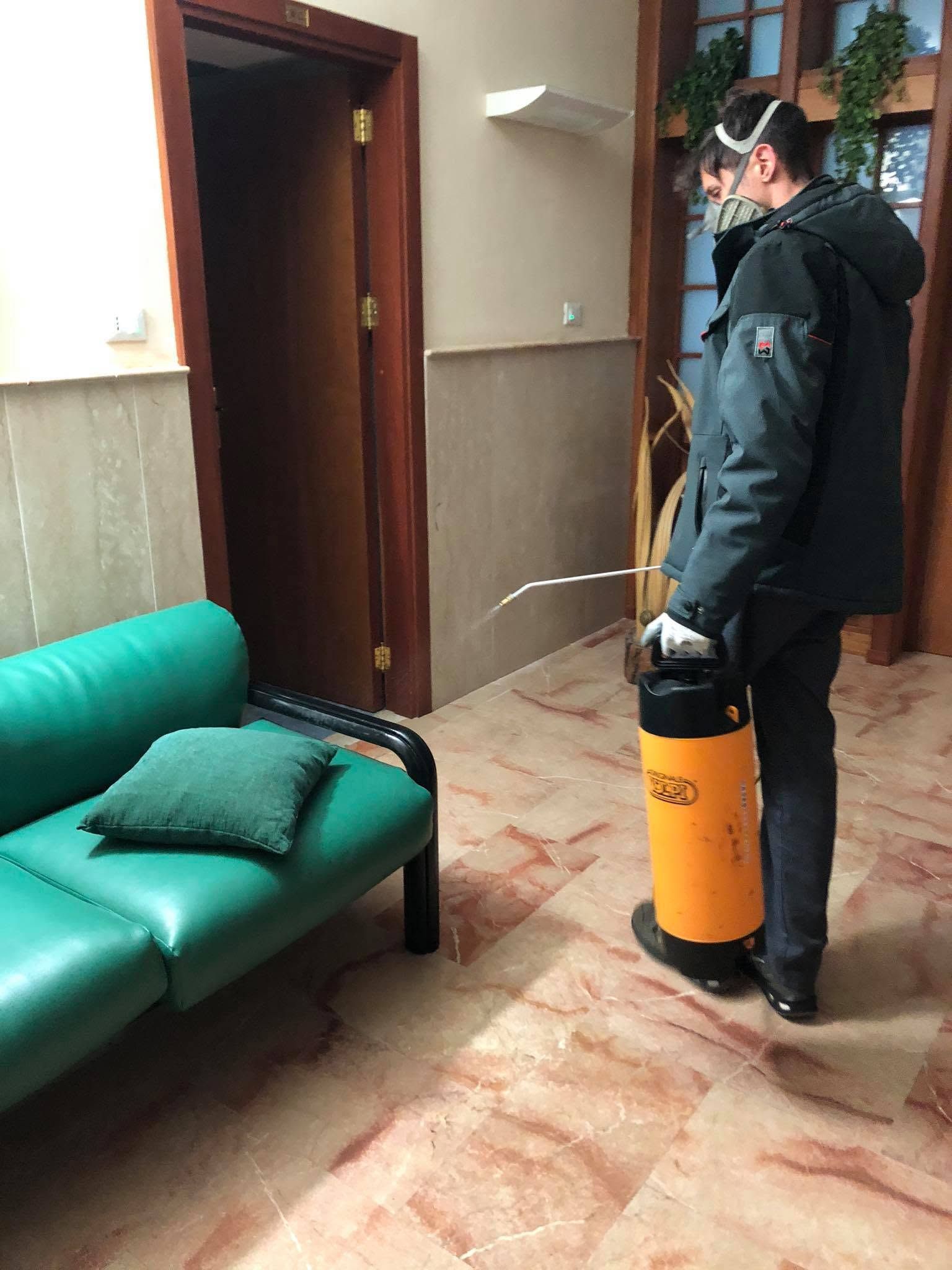 Una persona vestita di scuro usa uno spruzzatore arancione in un corridoio, accanto a un divano verde.