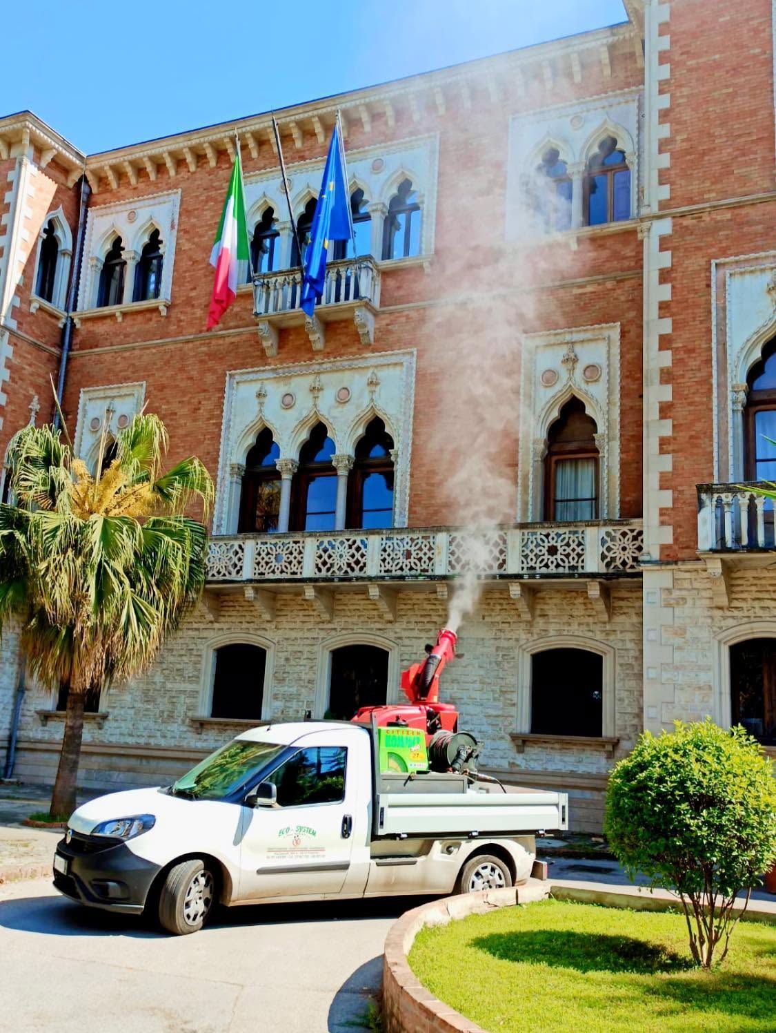 Un pick-up bianco con un operaio che aziona una piattaforma elevatrice rossa davanti a un edificio in mattoni con bandiere e una colonna di fumo