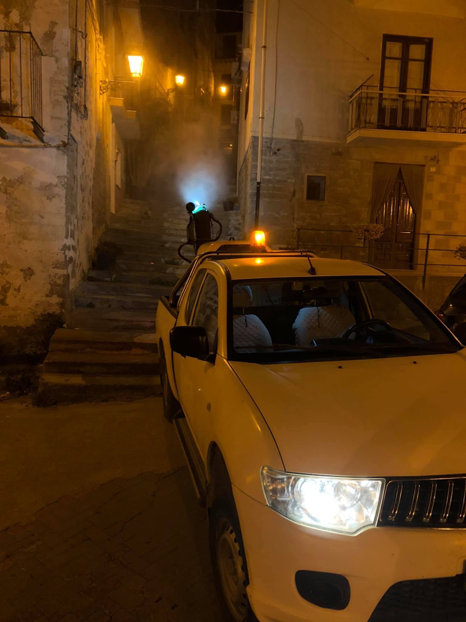 Auto bianca con i fari accesi di notte in una strada stretta; una persona è in piedi vicino a un muro illuminato da una luce blu.