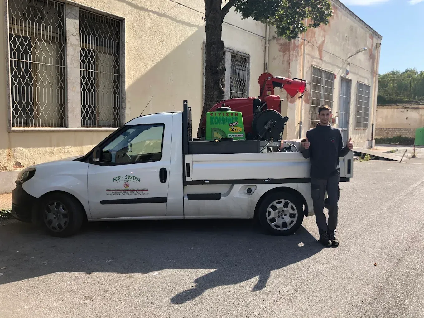 Un pick-up bianco carico di attrezzature è parcheggiato vicino a un edificio beige, con un uomo in piedi accanto.