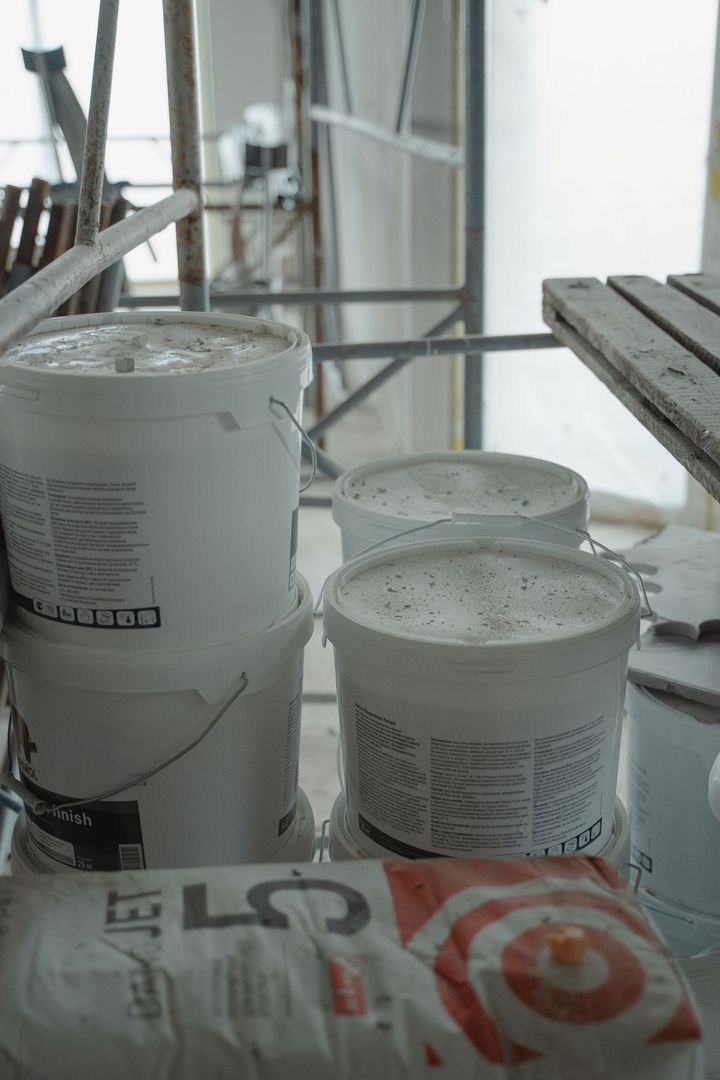 Two white paint buckets on a scaffold platform near a window with construction materials.