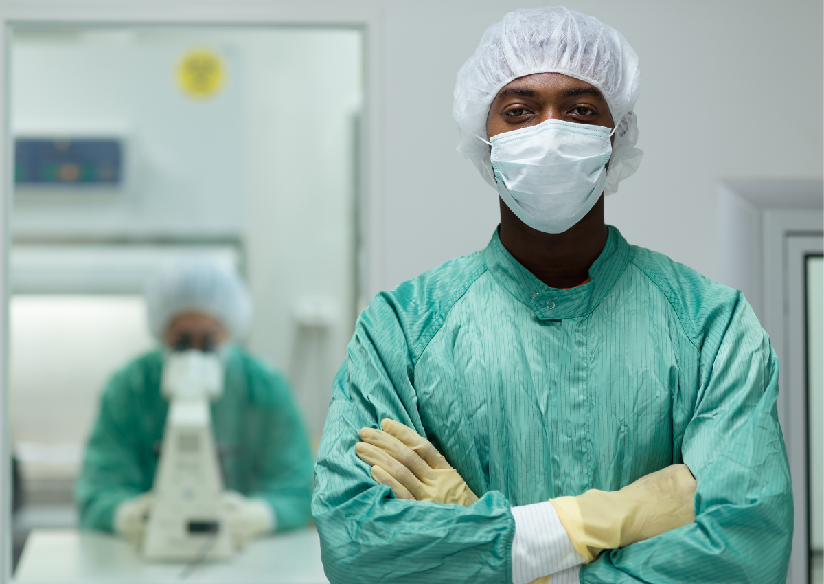 Un homme en combinaison de protection verte, masque et charlotte, les bras croisés dans un laboratoire.